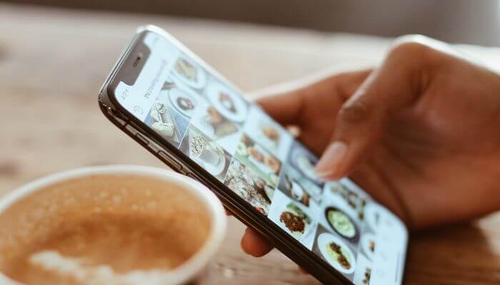 Foto de uma pessoa segurando um smartphone, explorando o feed do Instagram, mostrando várias imagens de comida.
