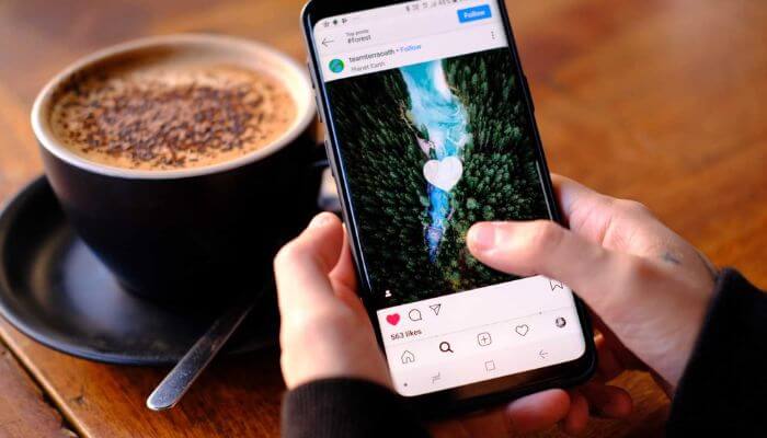 Foto de uma pessoa segurando um smartphone, interagindo com uma postagem no Instagram, mostrando um coração de curtida sobre uma imagem de natureza.