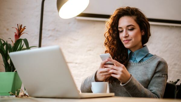 Mulher concentrada usando o celular enquanto trabalha em um notebook em um ambiente aconchegante.