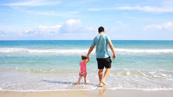 Pai e filha de mãos dadas caminhando na praia, com o mar ao fundo, exemplo de foto com legenda foto filha