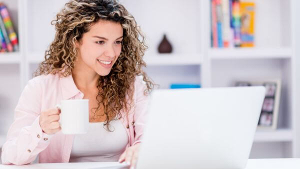 Mulher sorrindo enquanto trabalha em seu laptop e toma uma xícara de café. Overposting

