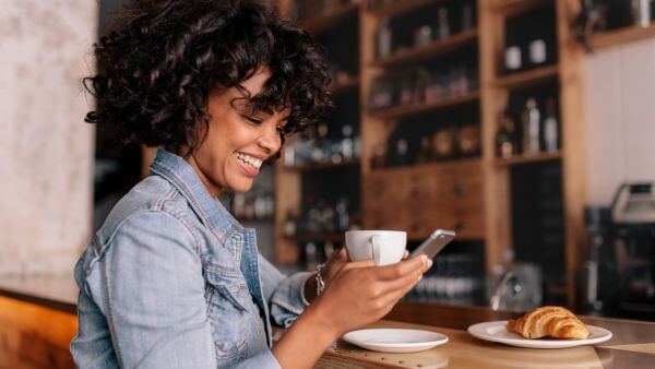 Mulher sorrindo enquanto toma café e usa o celular em uma cafeteria.