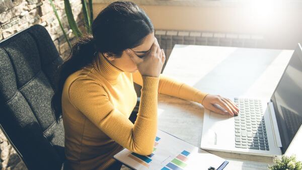 Mulher com expressão de cansaço, trabalhando em um laptop com gráficos ao lado. Overposting

