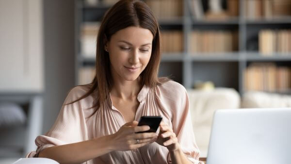 Mulher de cabelo liso, sentada em um ambiente aconchegante, segurando um celular buscando legenda photo dump e com um notebook aberto na mesa. Ao fundo, estante de livros. 