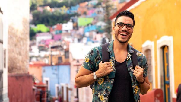 Jovem turista sorridente em uma rua colorida com casas vibrantes ao fundo. Perfeito para ilustrar um photo dump com momentos de viagem.